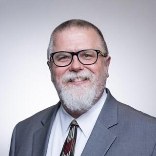 A headshot of a man in blue suit, white shirt and patterned tie with a beard and glasses.