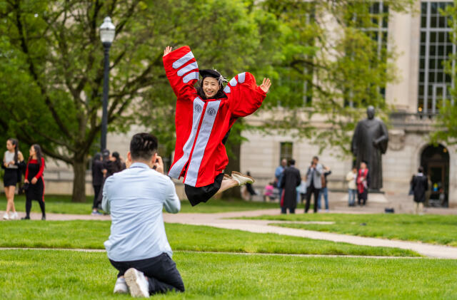 Student in red and gray Phd gown jumping in the air as another person kneels and takes a picture.