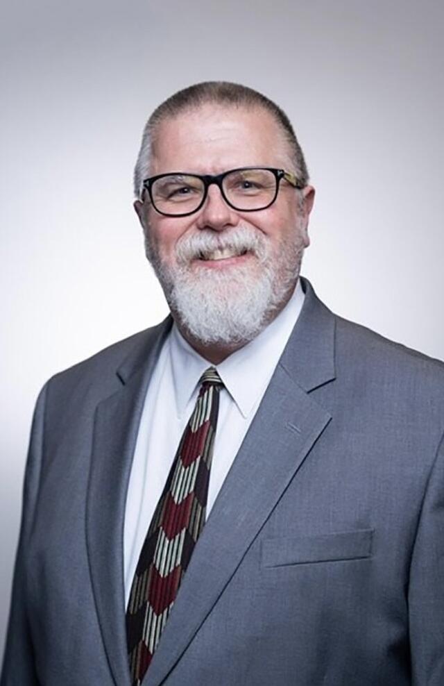 A headshot of a man in blue suit, white shirt and patterned tie with a beard and glasses.