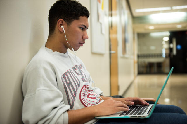 Man in OSU sweatshirt sitting in hallyway using laptop and headphones