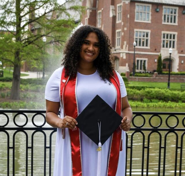Woman in a white dress holding a graduation cap outside.