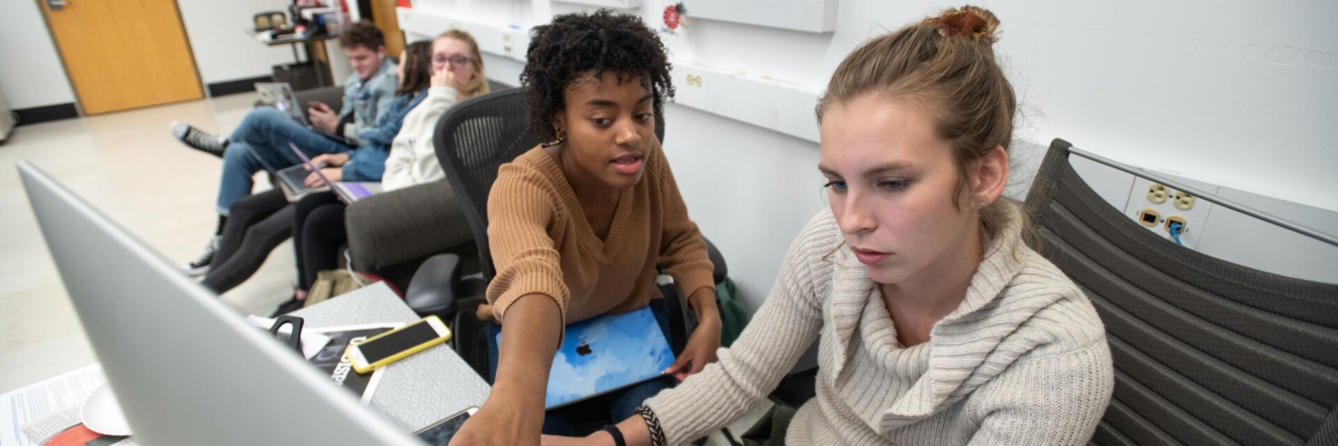 One woman sitting at a desk working with another woman pointing to the the computer screen.